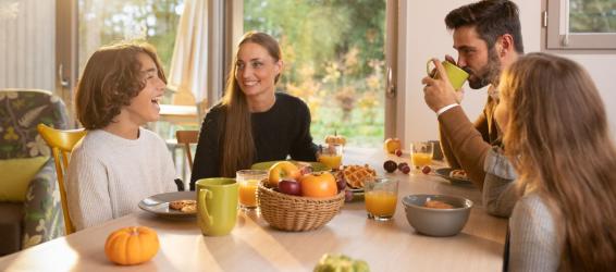 Famille réunie autour d’une table pour le petit-déjeuner, échangeant et souriant dans une pièce lumineuse avec vue sur la nature, fruits et boissons disposés sur la table.